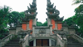 A traditional Balinese temple entrance with intricate stone carvings, surrounded by lush greenery. The structure features ornate details and a symmetrical design, typical of Balinese architecture video