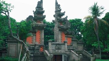 A traditional Balinese temple entrance with intricate stone carvings, surrounded by lush greenery and palm trees. The structure features red brick accents and is set against a cloudy sky video