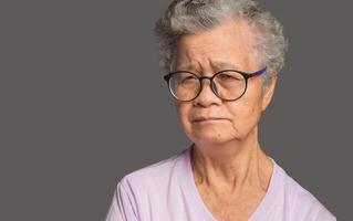 An elderly Asian woman is looking at the camera with a serious while standing on a gray background photo