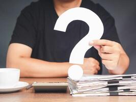 A young man in a black casual is holding a white question mark symbol while sitting at the table photo