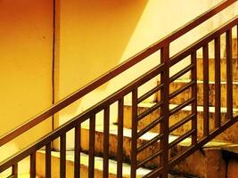 View of a geometric abstract image of a staircase with a yellow wall in the background. The railing is made of metal and is painted gold photo