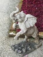 Grey elephant statue is standing on a stone path. The elephant is looking up. The statue is surrounded by red flowers, giving the scene a lively and colorful atmosphere photo