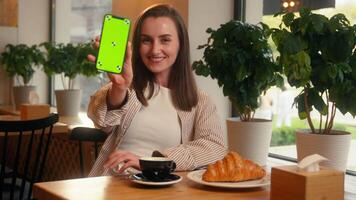 Smiling happy woman girl cafe visitor client in cafeteria restaurant female holding mobile phone showing green screen smartphone display mock up chroma key screen website menu order smile at camera video