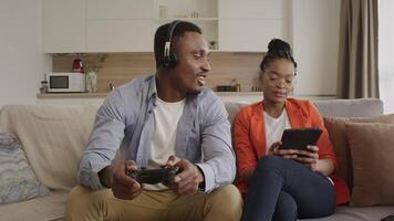 Happy african american couple chilling at home surfing the web on tablet and playing console games video
