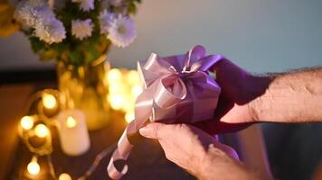 Male hands holding a gift box with a purple ribbon on a table with flowers, candles, and string lights, creating a romantic atmosphere Valentine's Day video