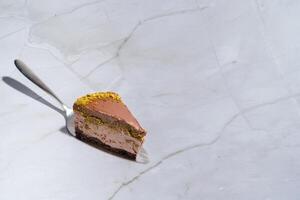 Slice of Dubai chocolate cheesecake with pistachio paste and kataifi dough covered in chocolate ganache on a cake spatula. Marble background with copy space. Indulgence, gourmet desserts, culinary photo