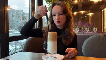 Woman guest stirs creamy coffee with spoon in coffeeshop lit by sunlight closeup. Lady orders caffeine drink with cake at rustic wooden counter. Morning vibes video