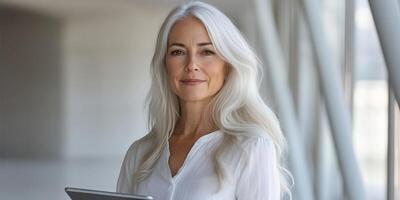 Professional woman with long gray hair holding a tablet in a modern office setting during the daytime photo