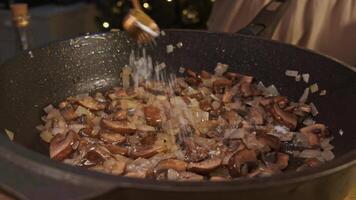Dark shiitake mushrooms are fried in pan with oil and sprinkled with salt, using measuring spoon to avoid over-salting. mushrooms are then mixed. video