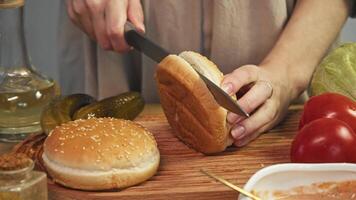 Close-up of housewife using sharp knife to cut sesame burger bun in half, surrounded by vegetables and other ingredients on cutting board. video