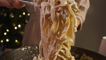 Chef in restaurant kitchen uses tongs to mix fettuccine pasta, mushrooms, and creamy white sauce on hot pan, creating an appetizing and nutritious dish. video