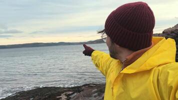 Young man with glasses and yellow raincoat stands against backdrop of ocean and rocky coast, dressed warmly on cold shores sea. He is communicating via call, sharing his impressions from trip to Arctic Circle. video