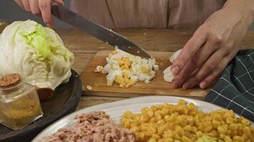 The chef in the kitchen is slicing a boiled egg with a sharp knife on a wooden chopping board surrounded by salad ingredients video