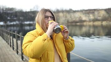 Tourist hipster woman in yellow winter jacket opening thermos, drinking coffee and relaxing at Zakrzowek lake, Krakow, Poland. Quarry under water. video