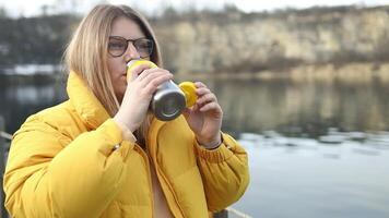 Tourist hipster woman in yellow winter jacket opening thermos, drinking coffee and relaxing at Zakrzowek lake, Krakow, Poland. Quarry under water. video