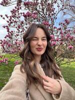 Caucasian woman with air touch hair coloring winks and taking a selfie near blooming magnolia tree photo