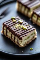 A slice of soft, crumbly pistachio halva is elegantly drizzled with dark chocolate. Served on a matte black plate, the dessert stands out against the softly blurred warm lights in the background photo