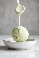 A beautifully sculpted sphere of pistachio gelato hovers elegantly above a porcelain plate, while smooth white chocolate flows gently down from the air, creating an enticing dessert photo