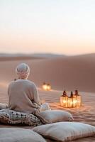 A traveler relaxes on luxurious Moroccan-style cushions in a private desert camp, enjoying the serene beauty of golden sand dunes and glowing lanterns as the sun sets photo