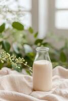 A glass milk bottle sits on a soft beige linen cloth, conveying a sense of freshness. Soft-focus green leaves in the background enhance the natural ambiance of this serene setting photo