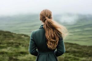 A woman stands in the vast Irish countryside, wearing a long green coat. She looks out over the rolling hills as soft mist rises in the distance, creating a serene atmosphere photo
