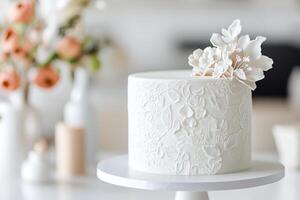 A smooth, white cake features delicate embossed floral details and is elegantly displayed on a stand. It complements a beautifully arranged setting of flowers, enhancing any celebration photo
