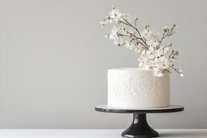 A smooth all-white cake features delicate embossed details, topped with branches of white flowers. It sits elegantly on a black cake stand against a neutral backdrop photo