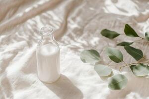 The glass milk bottle rests elegantly on a beige linen cloth, complemented by soft-focus green leaves in the background, creating a tranquil and fresh atmosphere photo