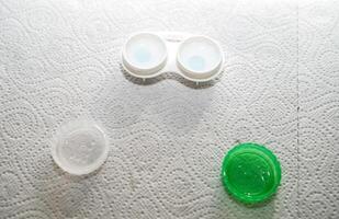 Container for storing lenses and contact lenses. The container is white with green, there are lenses in it. The table is covered with a paper towel for sterility at home. The container is open, you can see the lenses in it. photo