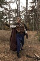 warrior girl with sword in hands in forest. blonde dressed in historical costume of warrior woman, with long cloak. warrior girl, raised sword, attacks enemy, looks at camera photo