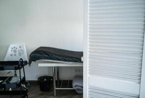 table for massage and other cosmetology services. covered with a blanket and a disposable sheet. next to it is a white chair. in the foreground is a white screen. photo
