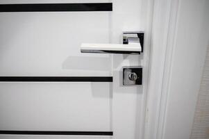 White door with silver handle and latch lock. On the door there are black narrow stripes parallel to the floor. The lock has a square shape. A close up of a door. High quality photo