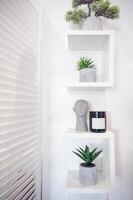 decorative shelf in white with artificial flowers in pots, black storage jar, and decorative sculpture head. next to the shelf is a white screen. vertical photo