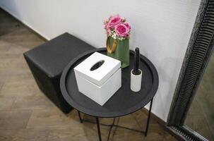 round black metal table next to it pouf. on the table are a box for napkins, decorative flowers, a brush for cleaning clothes. top view. on the floor are wood-like tiles. the walls are white photo