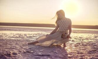 blonde girl sits in the middle of a salt lake. her dress flutters in the wind. a bright circle of the setting sun illuminates the shore covered with salt. she holds a bouquet in her hands, odessa photo