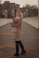 Blonde with a bouquet of pink tulips in the city in full growth. Girl with curls developing in the wind. She looks into the frame and smiles. Dressed in a beige classic coat. And a warm scarf photo