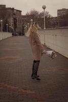 Blonde with a bouquet of pink tulips in the city in full growth. The girl dances with developing curls in the wind. She looks into the frame and smiles. She is dressed in a beige classic coat photo