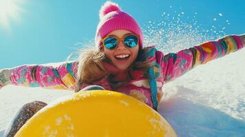 young girl sledding down a snowy hill. She is wearing a pink beanie, a colorful jacket, and sunglasses photo