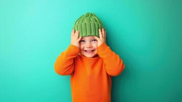 young child, probably around 2-3 years old, standing against a turquoise background. The child is wearing a bright orange sweater and a green knitted beanie photo