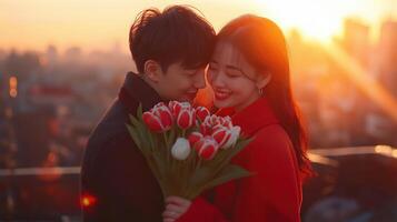 asian young couple embracing each other on a rooftop at sunset. The man is holding a bouquet of red and white tulips in his left hand and the woman is wearing a red coat photo