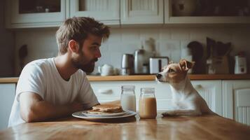 young man sitting at a kitchen table with a plate of pancakes in front of him. He is wearing a white t-shirt and has a beard. Next to him, there is a small dog photo