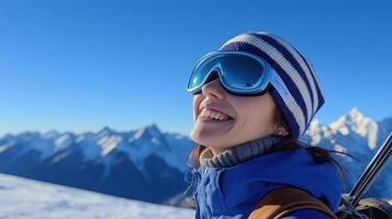 young woman wearing a blue and white striped beanie and ski goggles. She is standing on a snowy mountain with a clear blue sky in the background photo