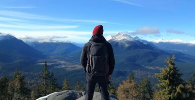 man standing on top of a mountain, looking out at a beautiful landscape. He is wearing a black jacket, a red beanie, and carrying a large black backpack photo