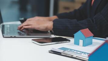 A man in a suit is typing on his laptop while holding a house model video