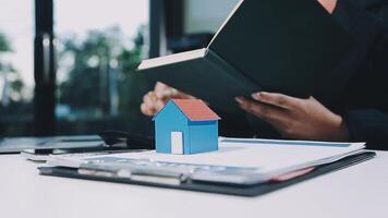 A real estate agent is holding a house model in front of a laptop video