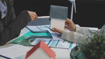 Two people sitting at a table with papers and a house model video