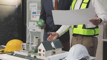 Two men in hard hats and safety vests are looking at a model of a building video