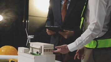 Two men in safety vests and hard hats are looking at a model of a building video
