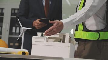 Two men in safety vests and hard hats are standing next to a model of a building video