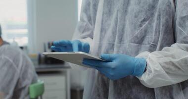 A male laboratory assistant in disposable medical clothing uses a digital tablet while working in the laboratory. Close-up of hands in rubber gloves typing on a tablet. 4k , red komodo video